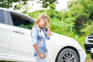 A woman cradles her head in her hands in front of a car damaged in a truck accident.