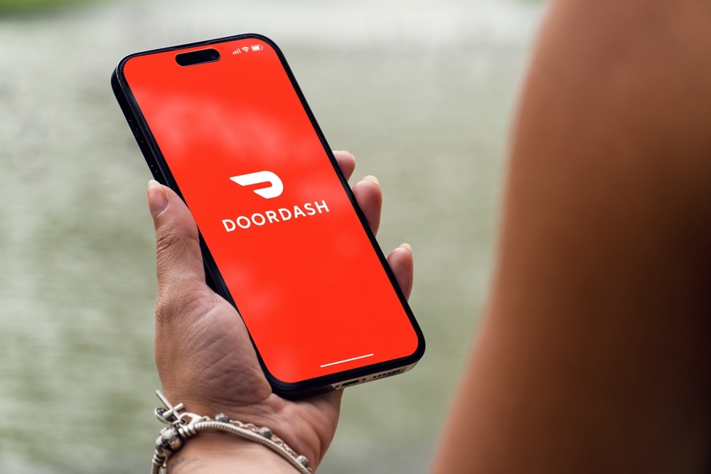 Girl in the park holding a smartphone iPhone 14 Pro with Doordash delivery app on the screen. Lake in the background. R